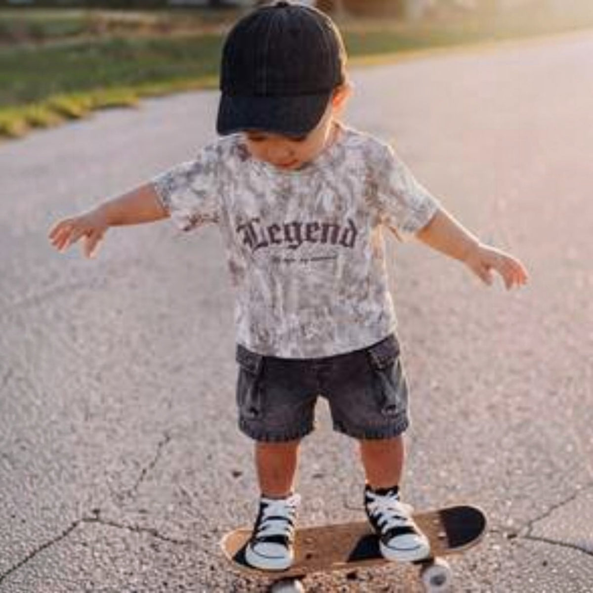 Babylove Nauru baby boy wearing a grey camo Legend t-shirt and black denim cargo shorts with a cap on a skateboard